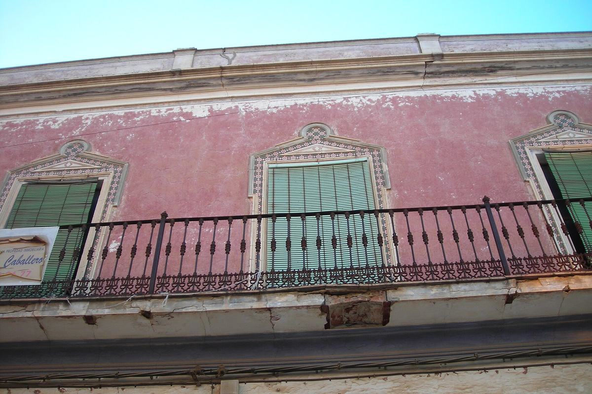 Fachada de una casa antigua con balcón de forja y un cartel de se vende